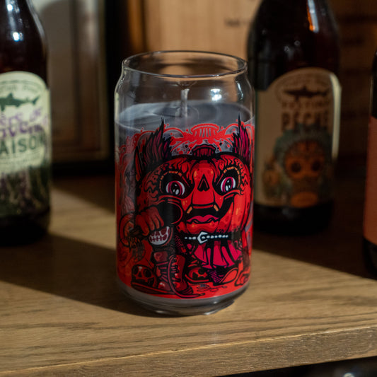 A clear glass featuring a bold red and black demon face sits on a shelf alongside brown beer bottles and the Punkin Can Glass Candle from Unwined Candles, LLC. Warm light highlights the candle, glass, and surrounding surfaces.