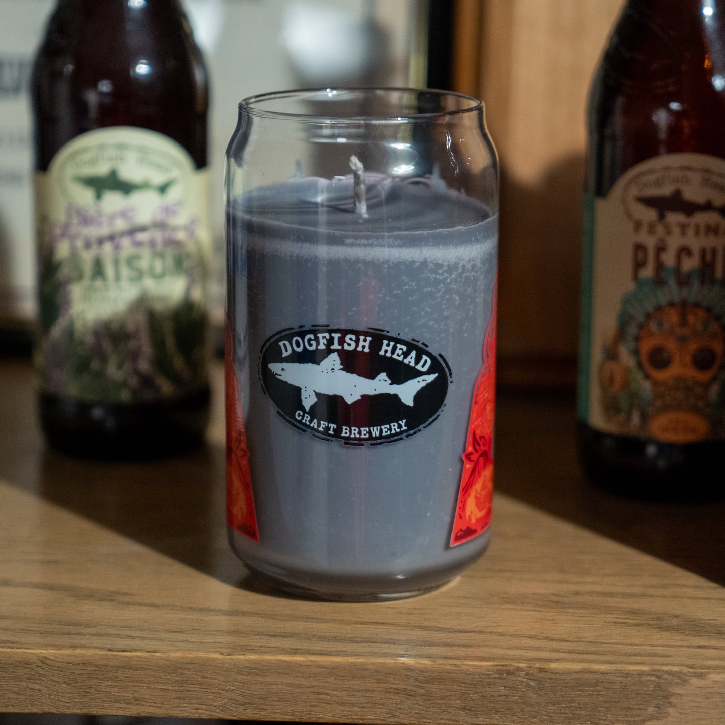 A gray Punkin Can Glass Candle by Unwined Candles, LLC sits on a wooden shelf between two Dogfish Head beers, making it perfect for enjoying the cozy pumpkin spice scent of this fall seasonal candle.