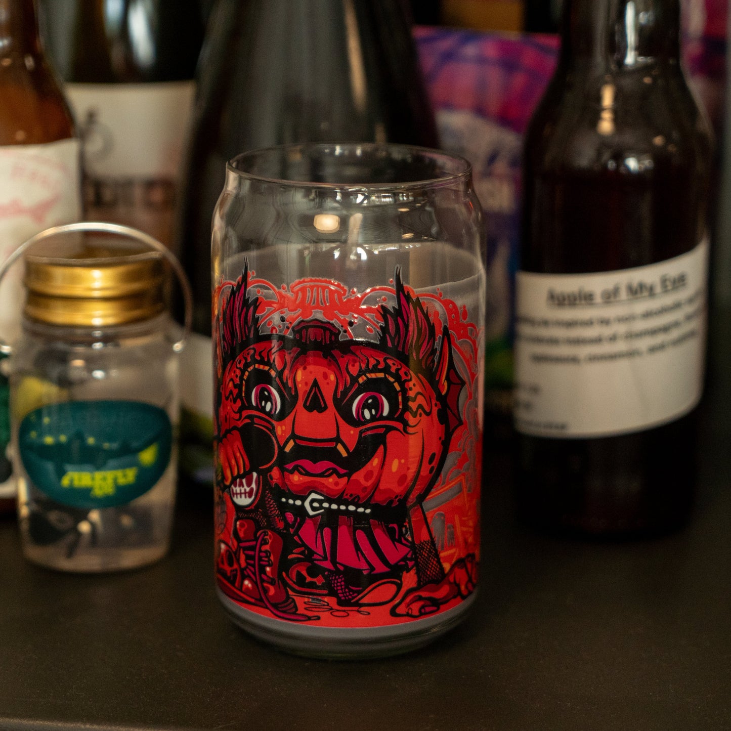 The Punkin Can Glass Candle by Unwined Candles, LLC features a vivid red and black pumpkin-headed design, displayed among bottles and jars—including a fall seasonal candle and the Apple of My Eye bottle.