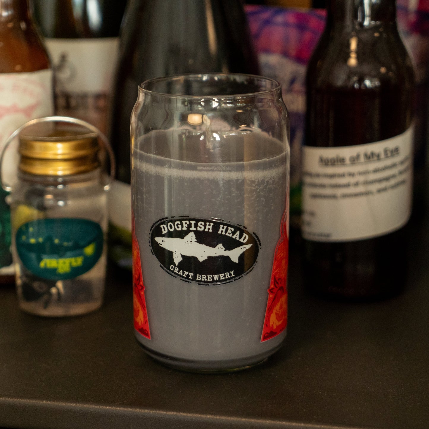 A glass labeled Dogfish Head Craft Brewery holds a cloudy, grayish-purple drink. Nearby is an Unwined Candles, LLC Punkin Can Glass Candle, filling the air with pumpkin spice amid assorted bottles and jars.