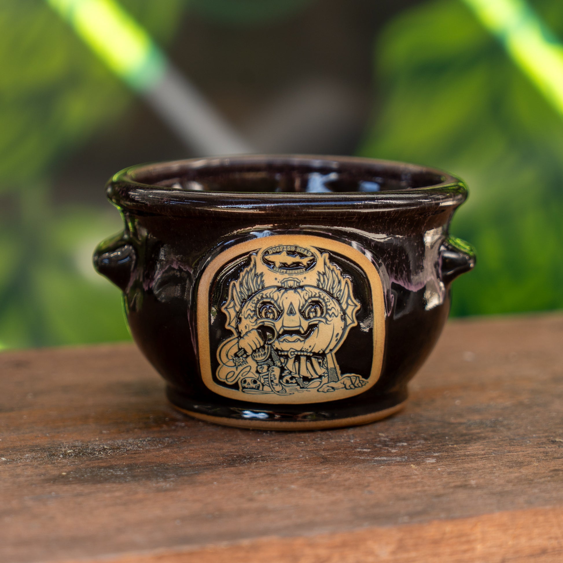 The Punkin Crock by Deneen Pottery is a dark ceramic piece with two handles, featuring Drew Millward's tan engraved face and crown design, displayed on wood with a soft green background.