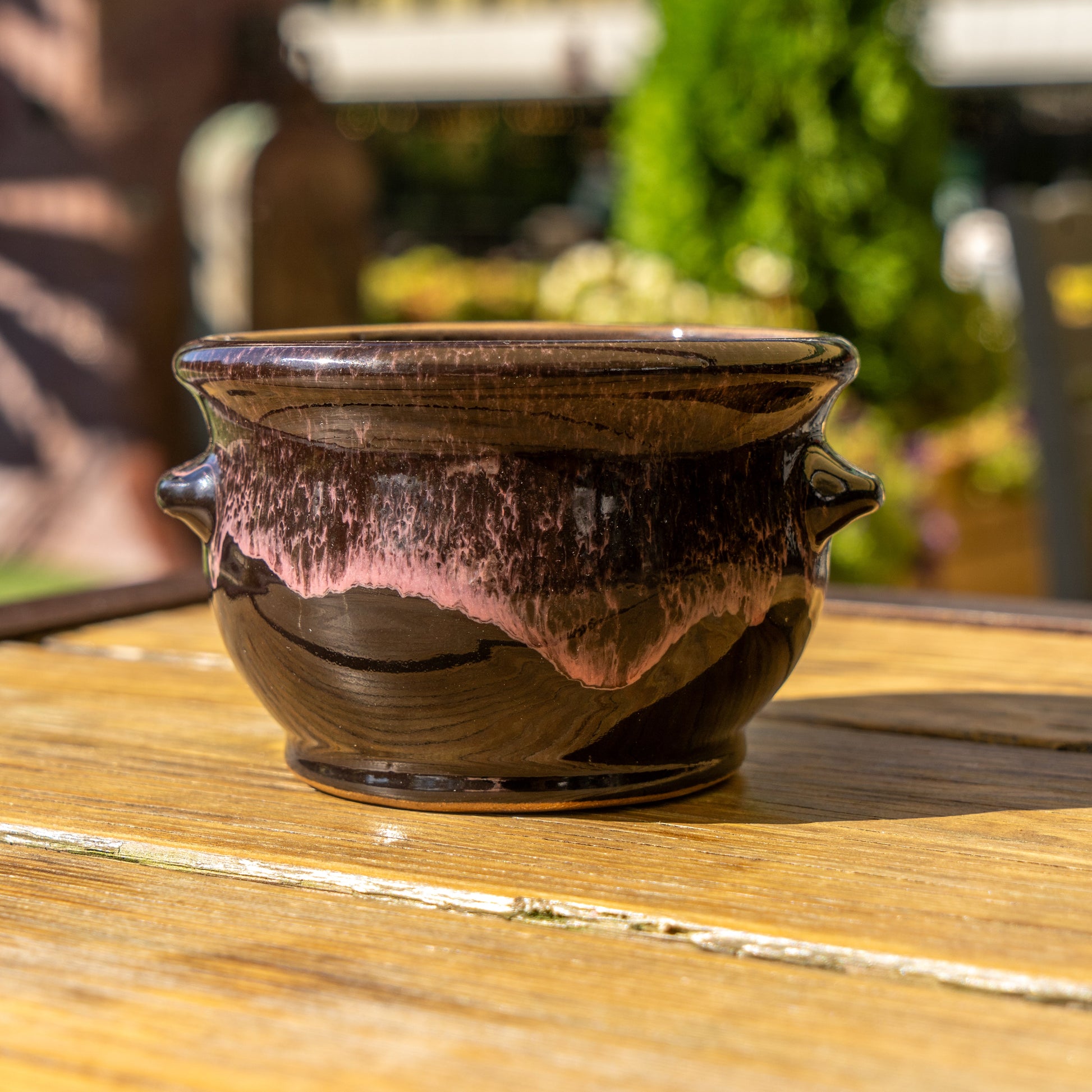 The Punkin Crock by Deneen Pottery—a glossy dark brown ceramic piece with pinkish glaze accents—rests on a wooden table outdoors, sunlight highlighting its details as greenery blurs in the background; ideal for seasonal décor.