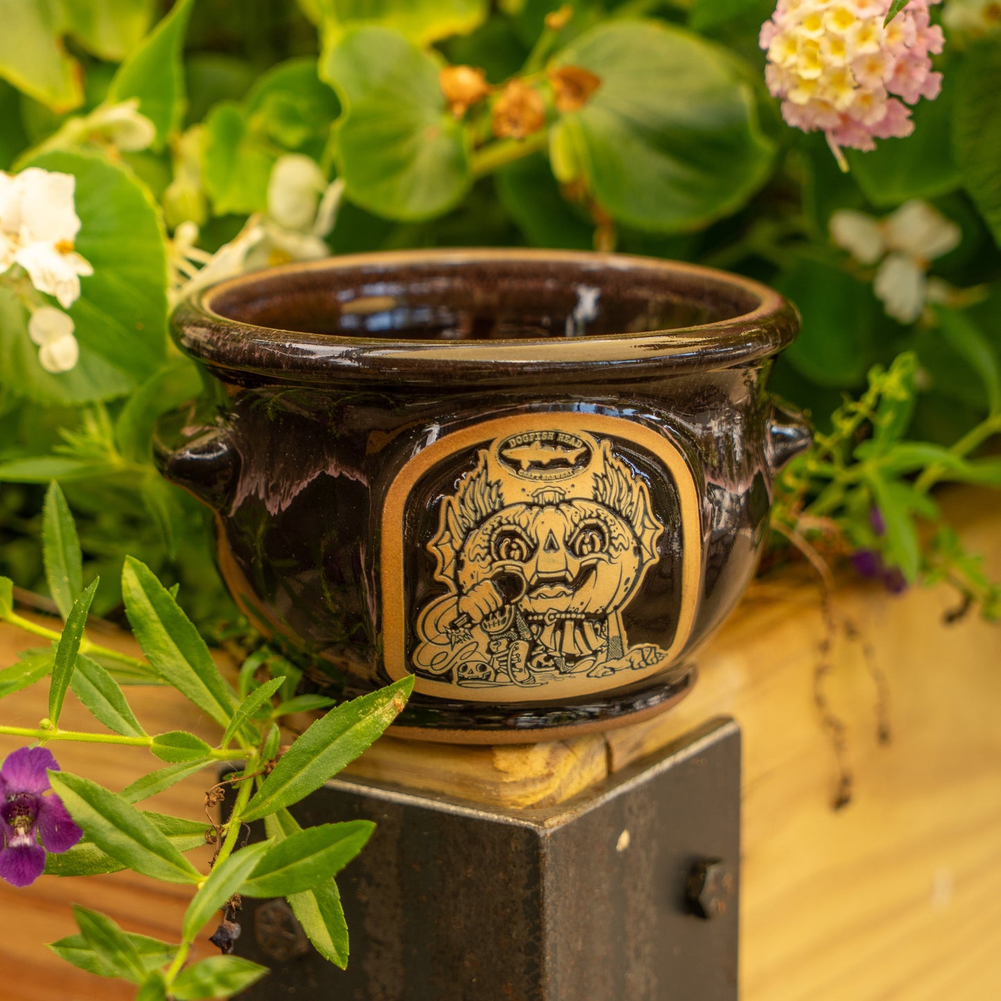 The Punkin Crock by Deneen Pottery, featuring a whimsical Drew Millward creature, rests on a wooden ledge amid lush green leaves and vibrant flowers in an outdoor garden.