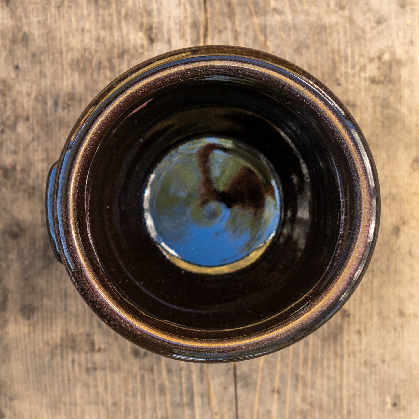 Top-down view of the Punkin Crock mug by Deneen Pottery—a dark, glossy ceramic piece featuring a subtle blue reflection inside—set on a weathered wooden surface, released as part of a special seasonal drop.
