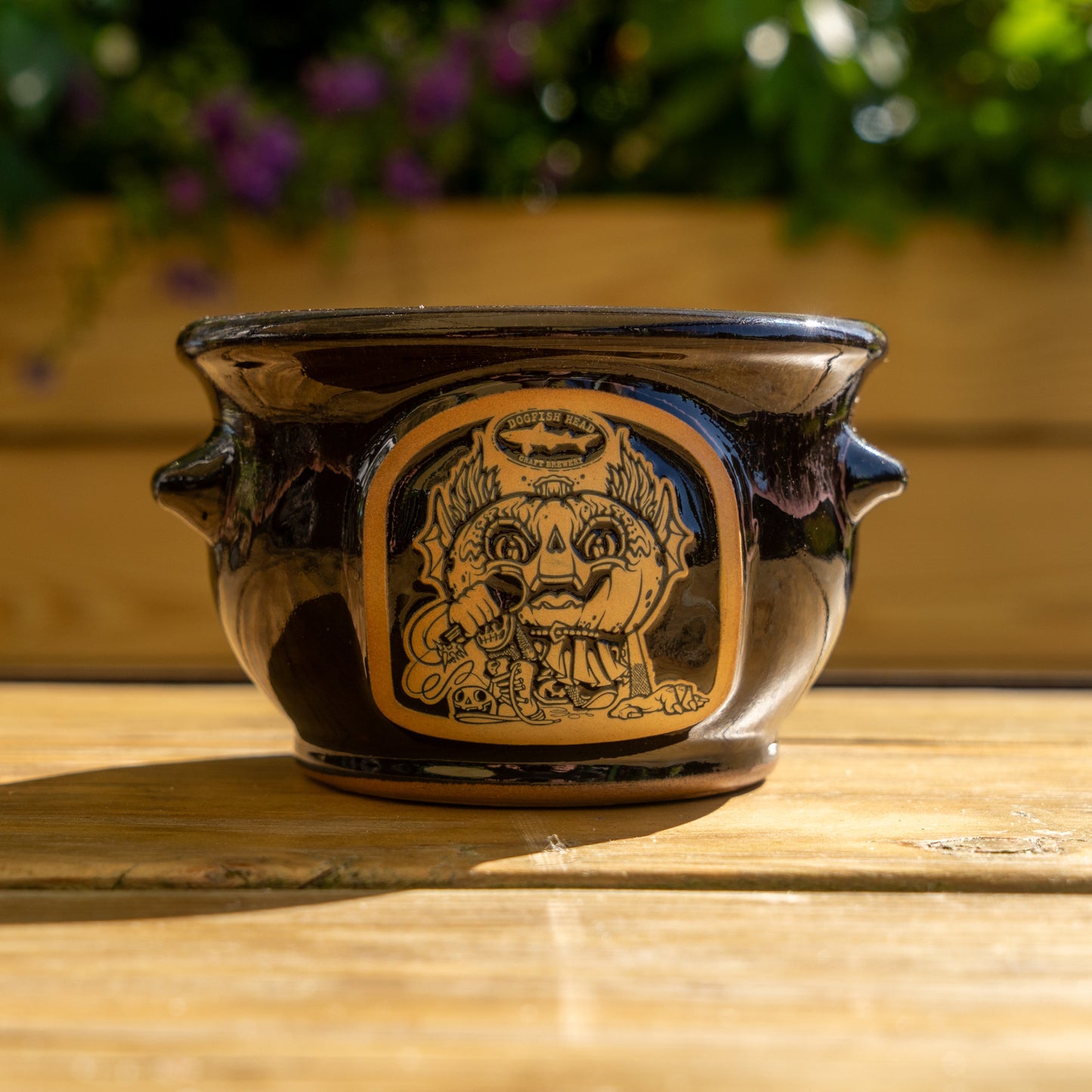 The Punkin Crock by Deneen Pottery is a black ceramic piece with two handles and a tan and black whimsical face illustration, resting on a sunlit wooden surface with greenery in the blurred background.