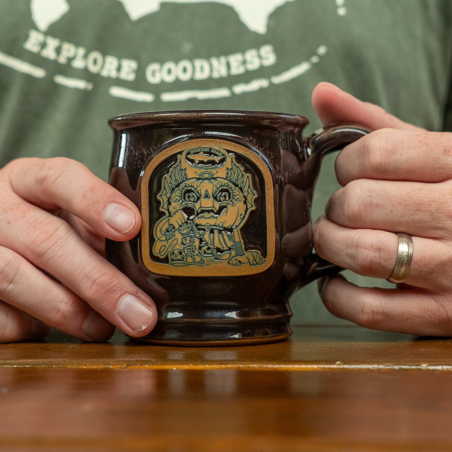 Close-up of a person holding the Deneen Pottery Punkin Mug, featuring artwork of two pumpkins and a cat. The person wears a ring and a green shirt with partially visible "EXPLORE GOODNESS" text.