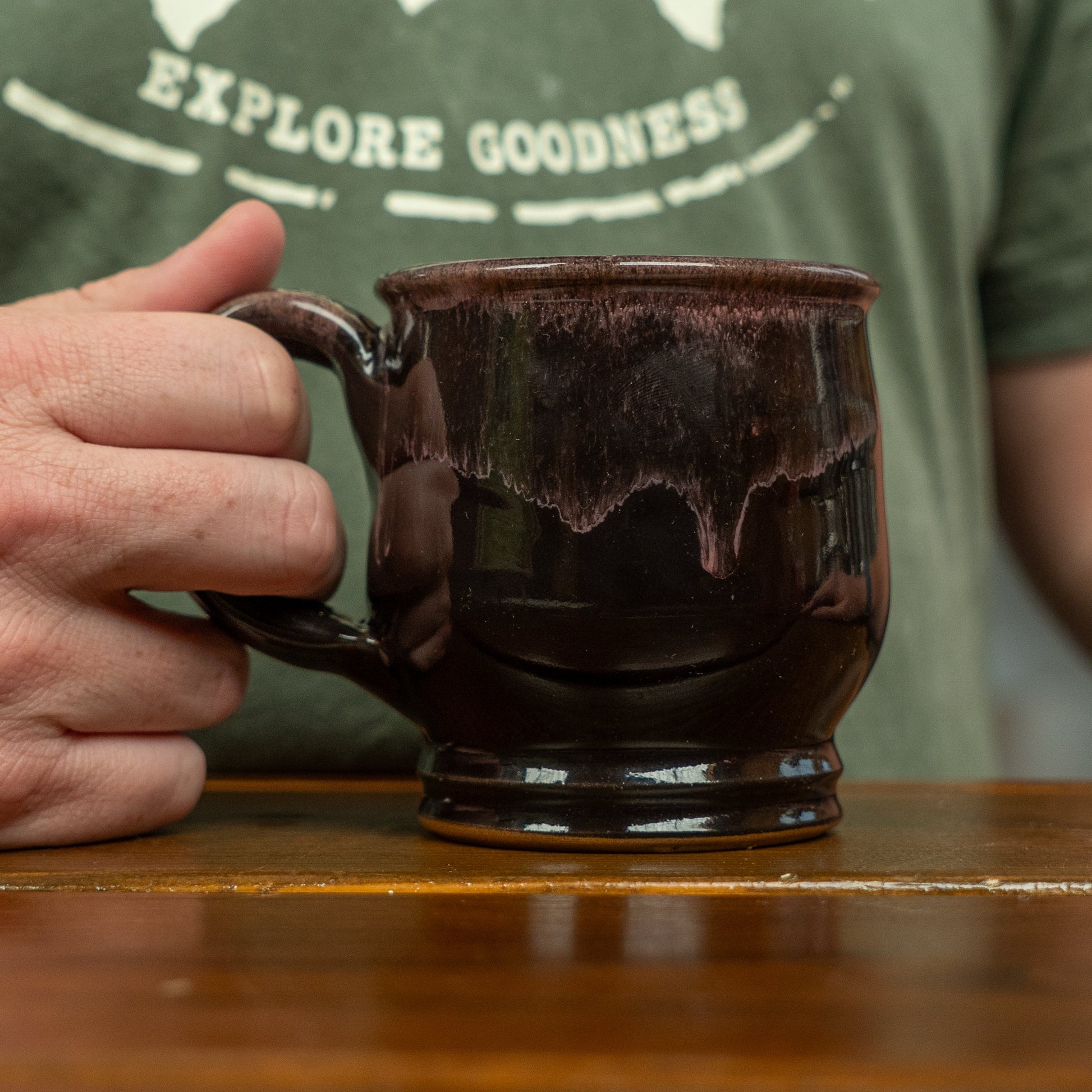 A person in a green shirt with partial white "EXPLORE GOODNESS" text holds a Deneen Pottery Punkin Mug by the handle, resting it on a wooden surface.