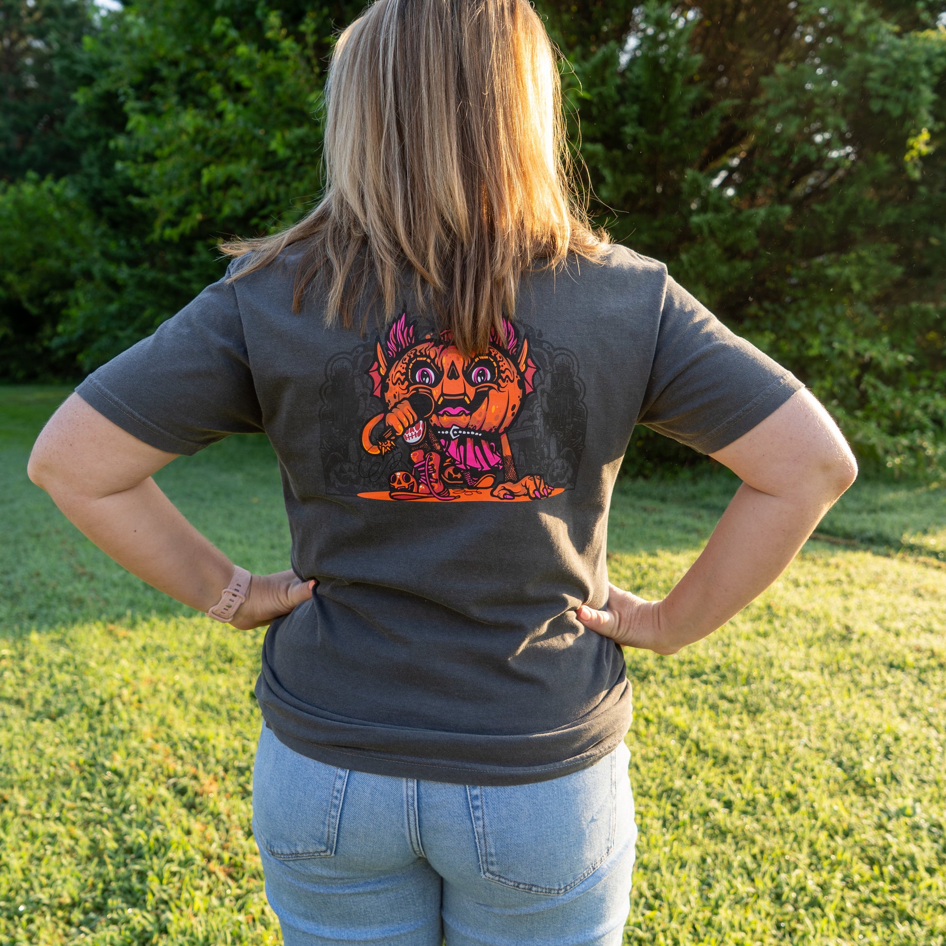 A person stands on grass wearing light blue jeans and a gray Punkin Short Sleeve Tee by GO WEST T-SHIRT COMPANY, featuring a colorful cartoon monster graphic on the back. Sunlight and trees enhance the bright, seasonal look.