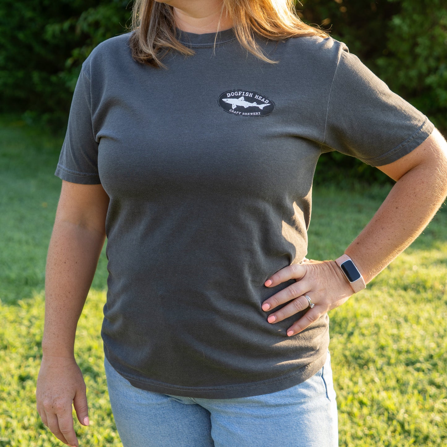 A woman outdoors on grass, hand on hip, wears a gray GO WEST T-SHIRT COMPANY Punkin Short Sleeve Tee reading “Dogfish Head Craft Brewery” and light blue jeans. She also has a smartwatch and ring; her face isn’t visible.