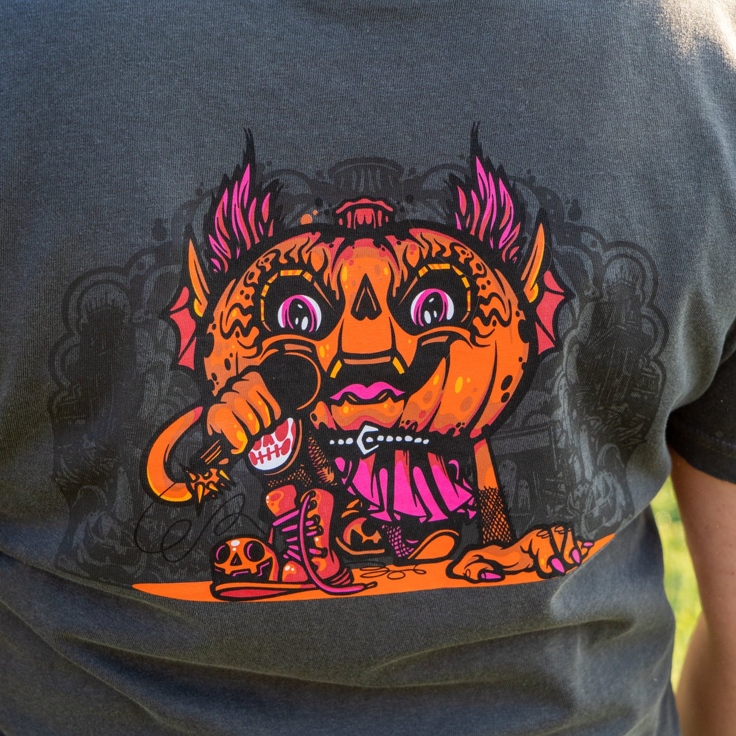 A close-up of the GO WEST T-SHIRT COMPANY's Punkin Short Sleeve Tee, featuring a vibrant cartoon pumpkin with bat wings and a mic, set against bold designs on a dark backdrop—your must-have seasonal shirt!.