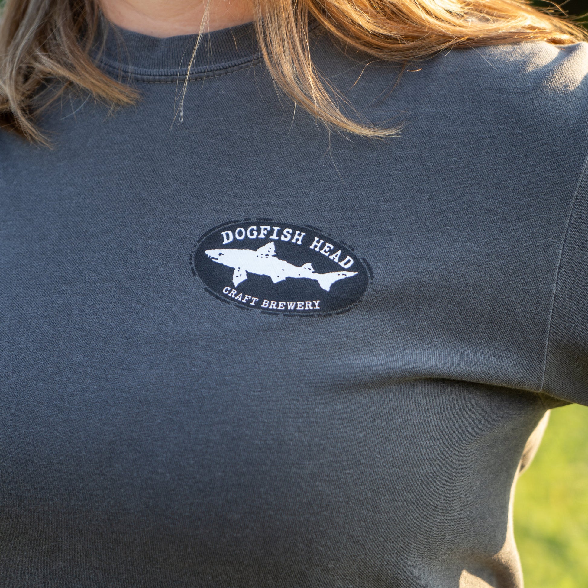 A person wearing the GO WEST T-SHIRT COMPANY Punkin Short Sleeve Tee, a garment-dyed dark gray shirt with a Dogfish Head Craft Brewery shark logo on the chest, stands outdoors with sunlight on their shoulder.