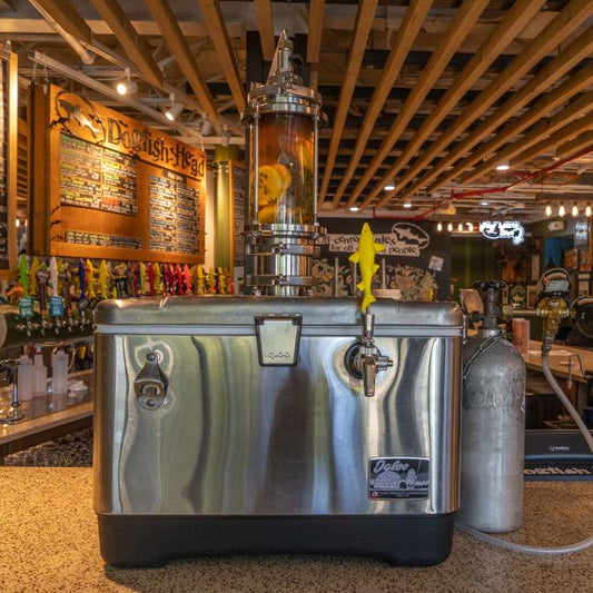 A silver cooler has been converted into a jockey box and sits on a bar counter. A stainless-steel draft tower with a clear cylinder containing citrus fruit slices rises from the top of the cooler. A yellow Dogfish Head shark tap handle is attached to the faucet on the front. A silver CO2 tank stands next to the cooler on the right. 