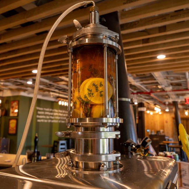 Close-up of a stainless steel draft tower rising from a jockey box. The tower features a clear cylinder filled with a dark liquid and slices of citrus fruit, possibly lemons or limes. A clear tube connects to the top of the cylinder. The background is blurred but shows a bar or restaurant interior with wooden ceiling beams and warm lighting.