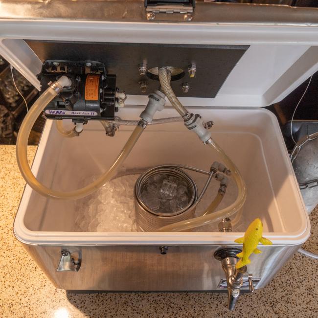 An open silver cooler reveals the inner workings of a jockey box. A small black pump is attached to the inside of the lid. A yellow Dogfish Head shark tap handle is visible on the outside faucet.