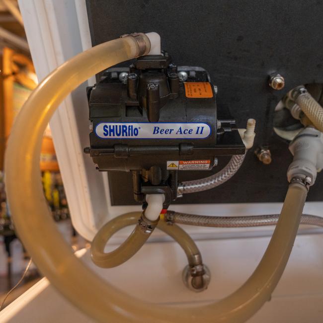 Close-up of a black Shurflo Beer Ace II pump attached to the inside of a jockey box lid. Clear beer lines with hose clamps connect to the pump. The inside of the white cooler and other tubing are partially visible. 
