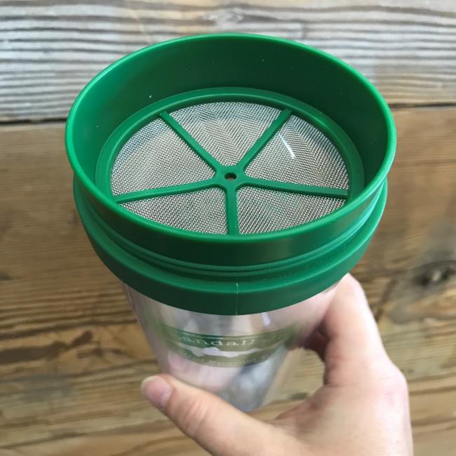 A hand holds the Randall Jr stainless steel tumbler by Arrasmith Promotions LLC, featuring a green lid and built-in fine mesh strainer, ideal for infusions. Wooden paneling forms the background.