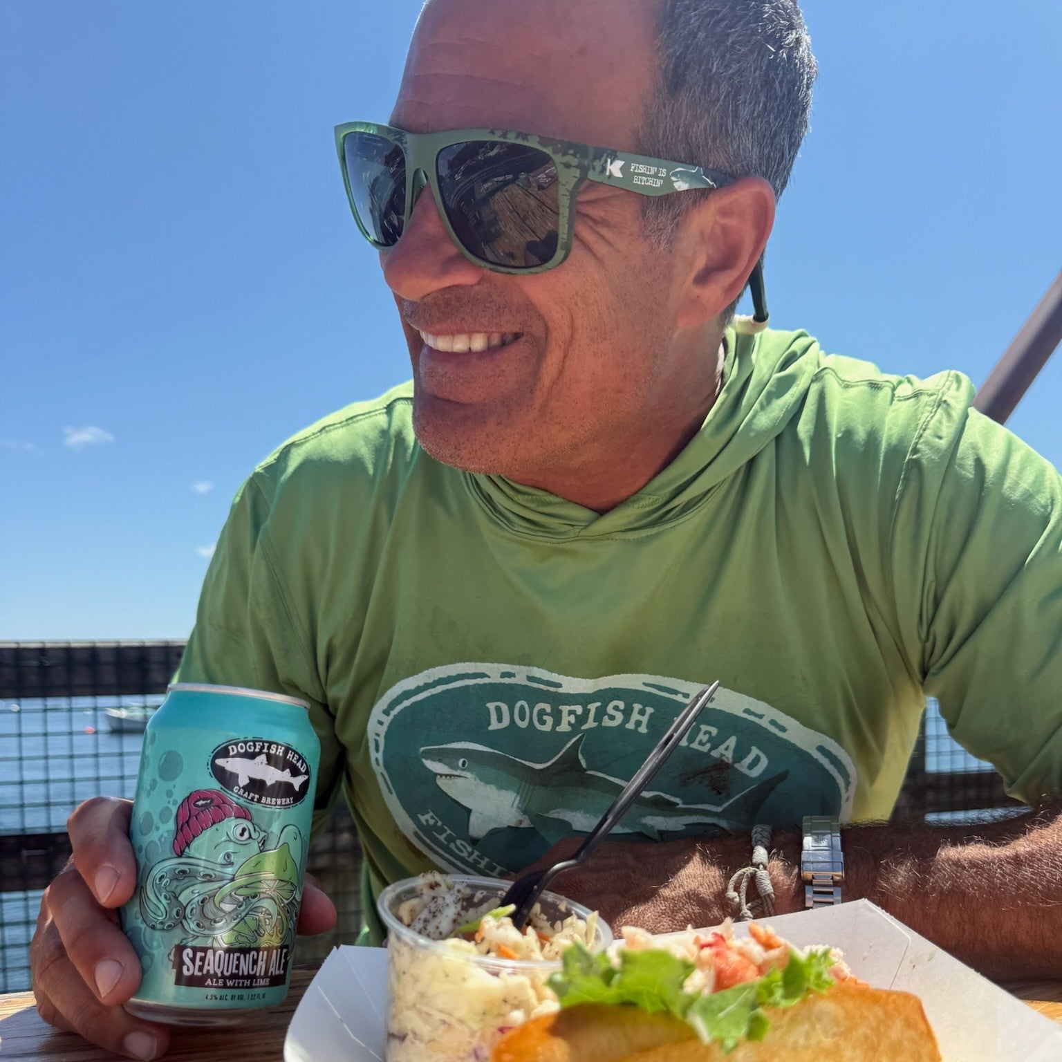 A man wearing green sunglasses and a green Dogfish Head t-shirt sits at an outdoor table holding a can of Dogfish next to a food tray with a fish taco and coleslaw.