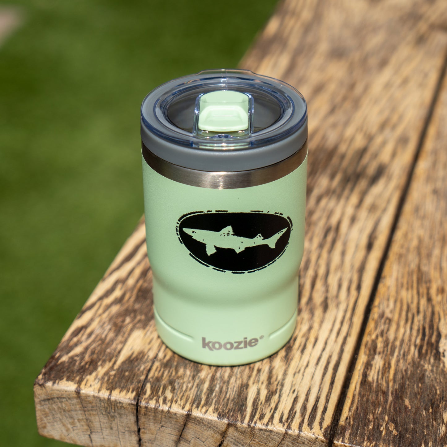 A Sage 12oz Koozie Can Insulator by Arrasmith Promotions LLC with a black shark graphic and clear lid sits on a wooden surface with grass in the background.