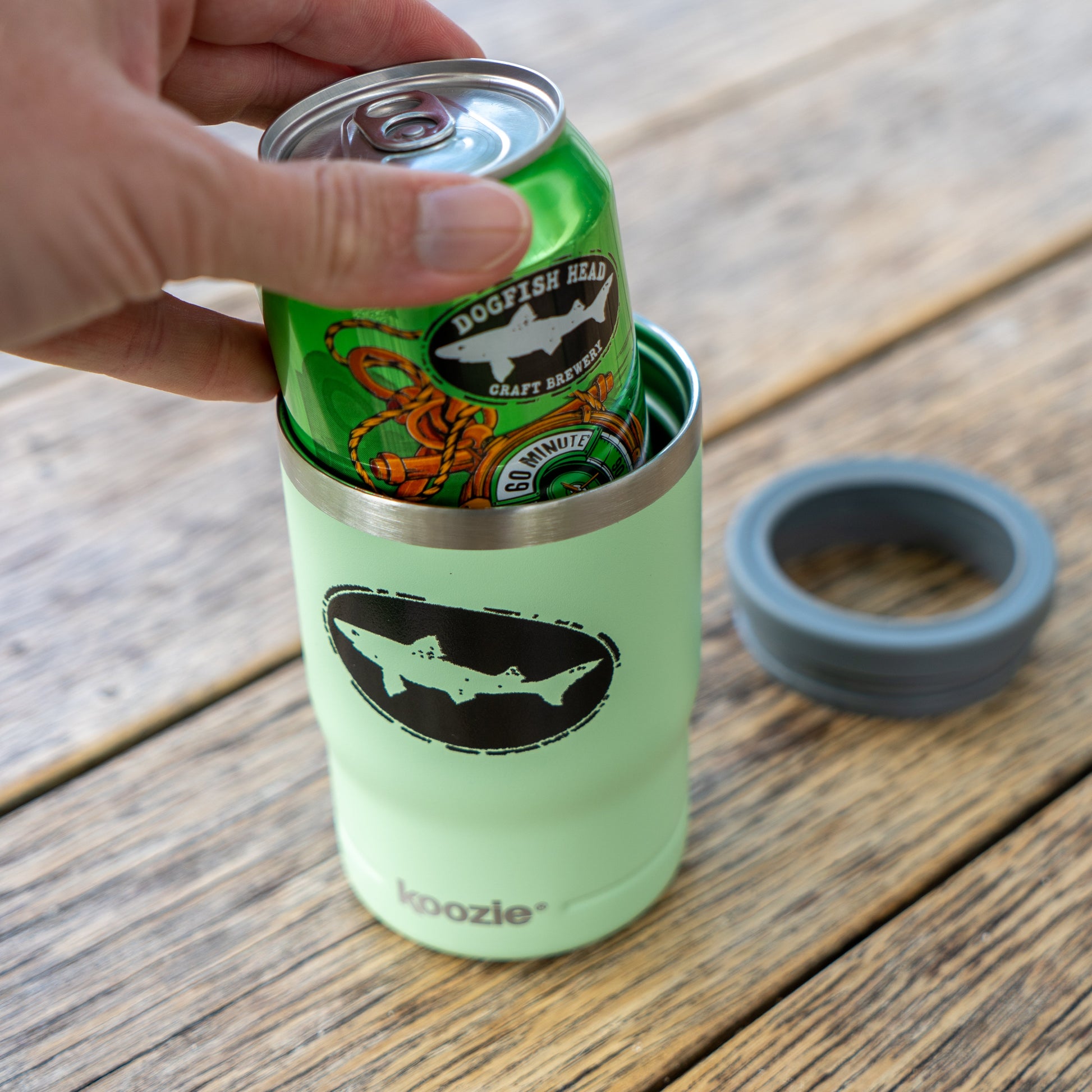A hand places a green Dogfish Head beer can into an Arrasmith Promotions LLC Sage 12oz Koozie Can Insulator on a wooden table. A gray lid sits beside the vacuum-insulated tumbler, ready to keep your drink cold.