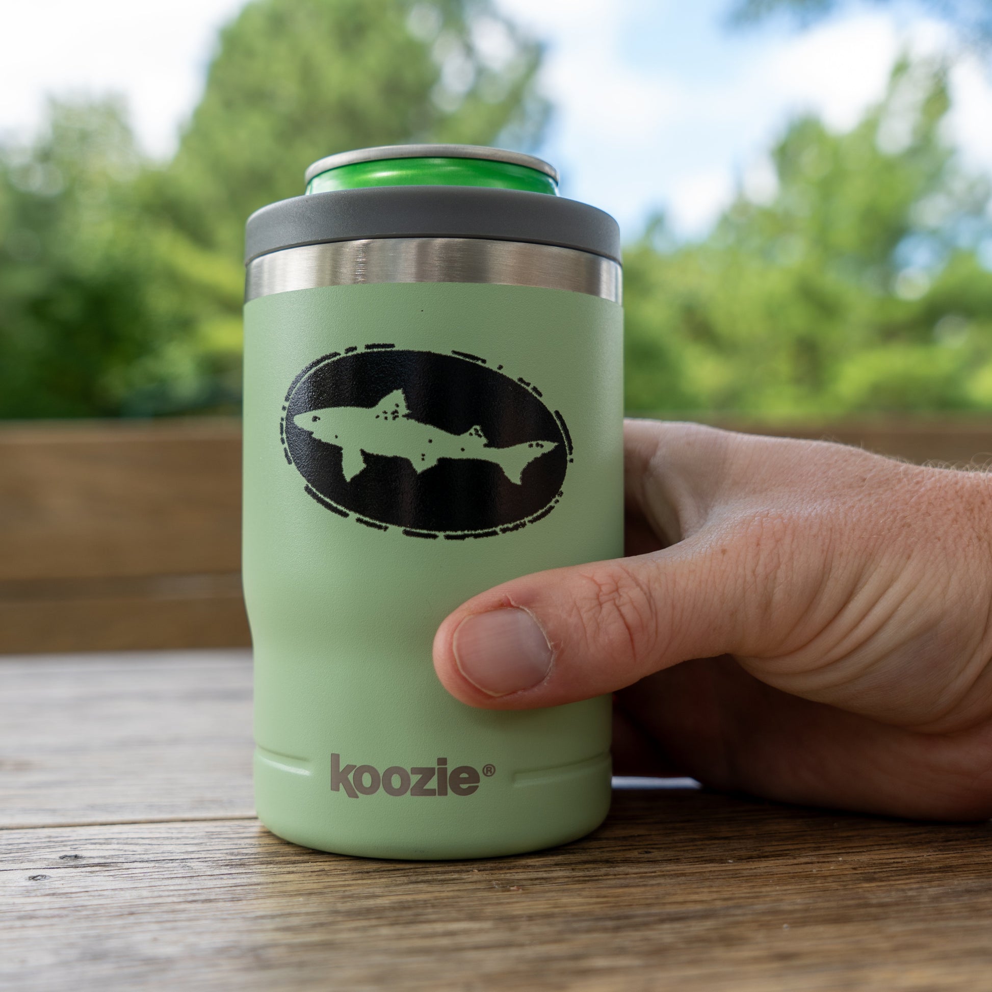 A hand holds a Sage 12oz Koozie Can Insulator by Arrasmith Promotions LLC, with a green can inside. The scene is outdoors, featuring a blurred wooden surface and green trees in the background.