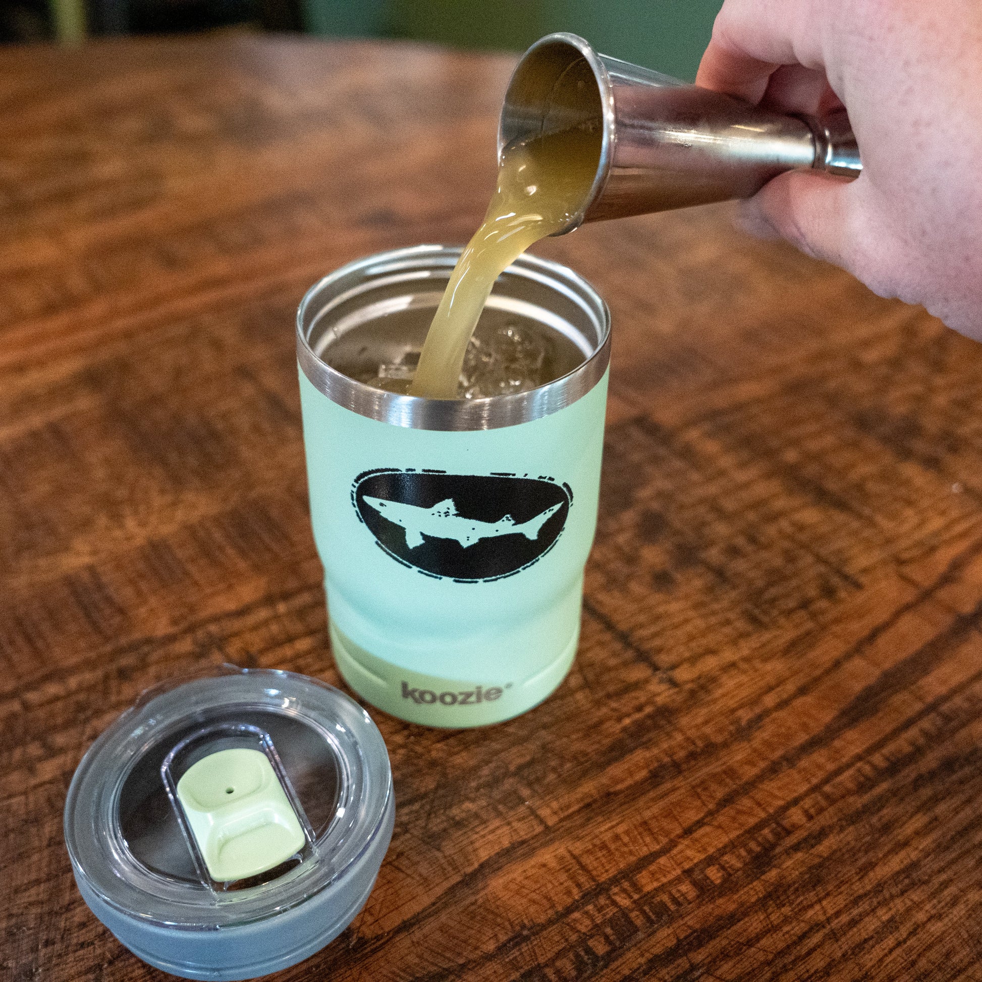 A hand pours a light brown liquid from a metal jigger into an Arrasmith Promotions LLC Sage 12oz Koozie Can Insulator, mint green with a shark logo, on a wooden table; the lid rests nearby.