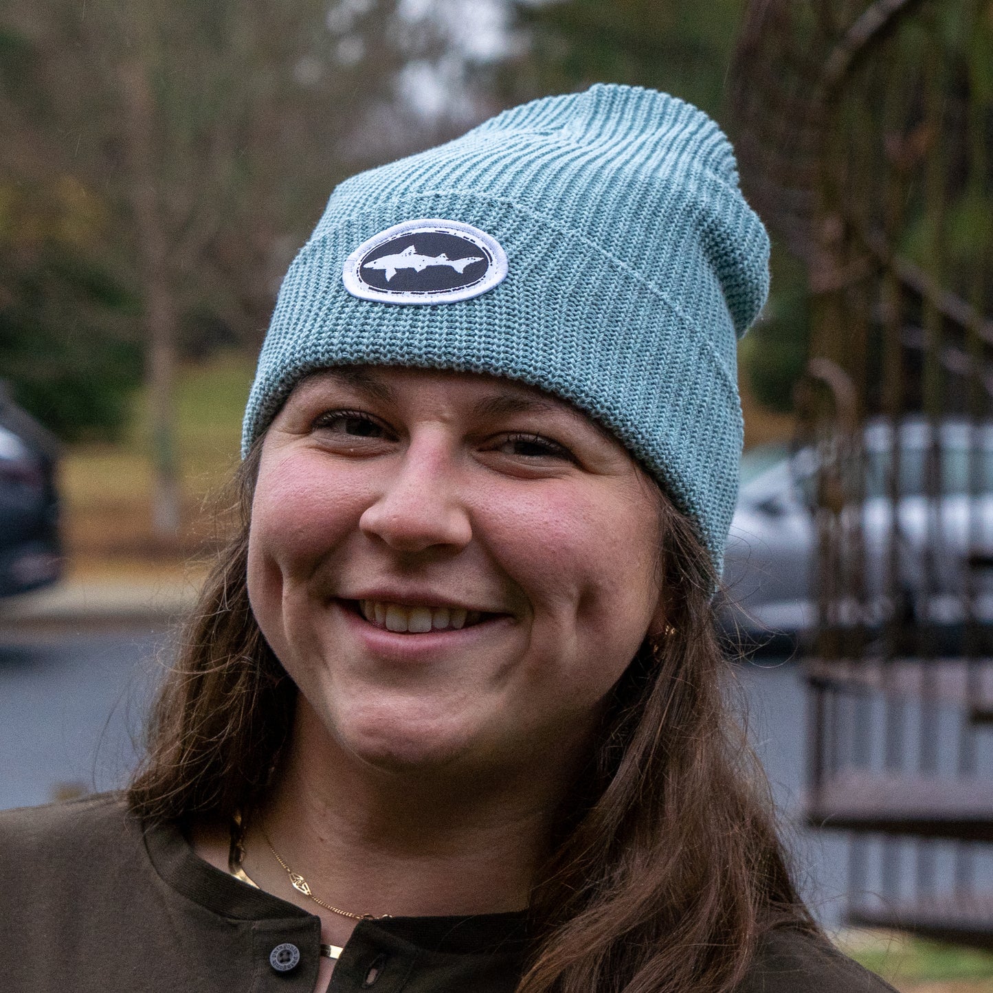 A smiling woman with long brown hair wears the Upstate Stock Seafoam Patch Beanie, featuring an off-center woven patch, while standing outdoors with blurred trees and cars in the background.