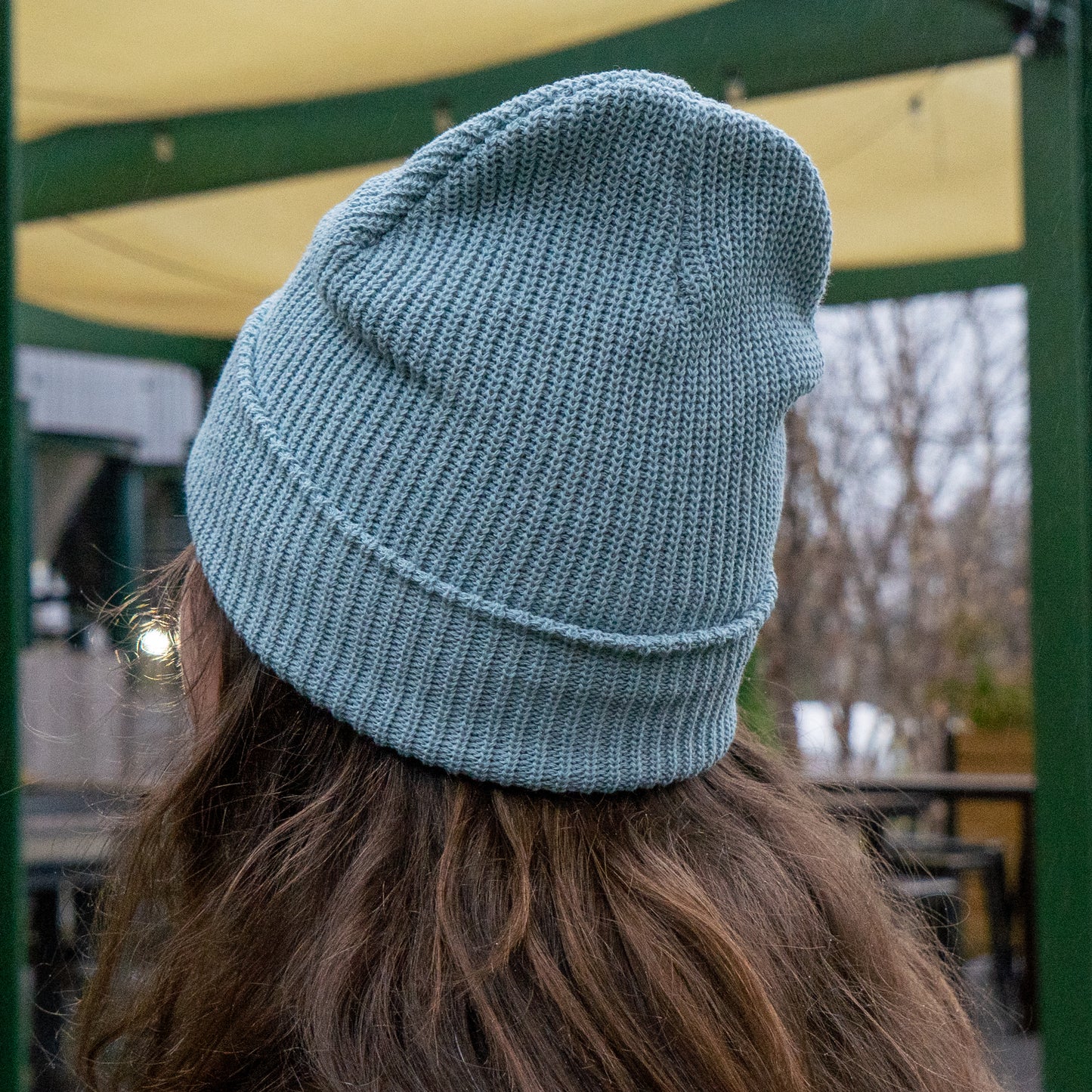 A person with long brown hair, seen from behind under a green and yellow canopy, wears the Upstate Stock Seafoam Patch Beanie—a light blue knit hat with a woven patch set off-center.