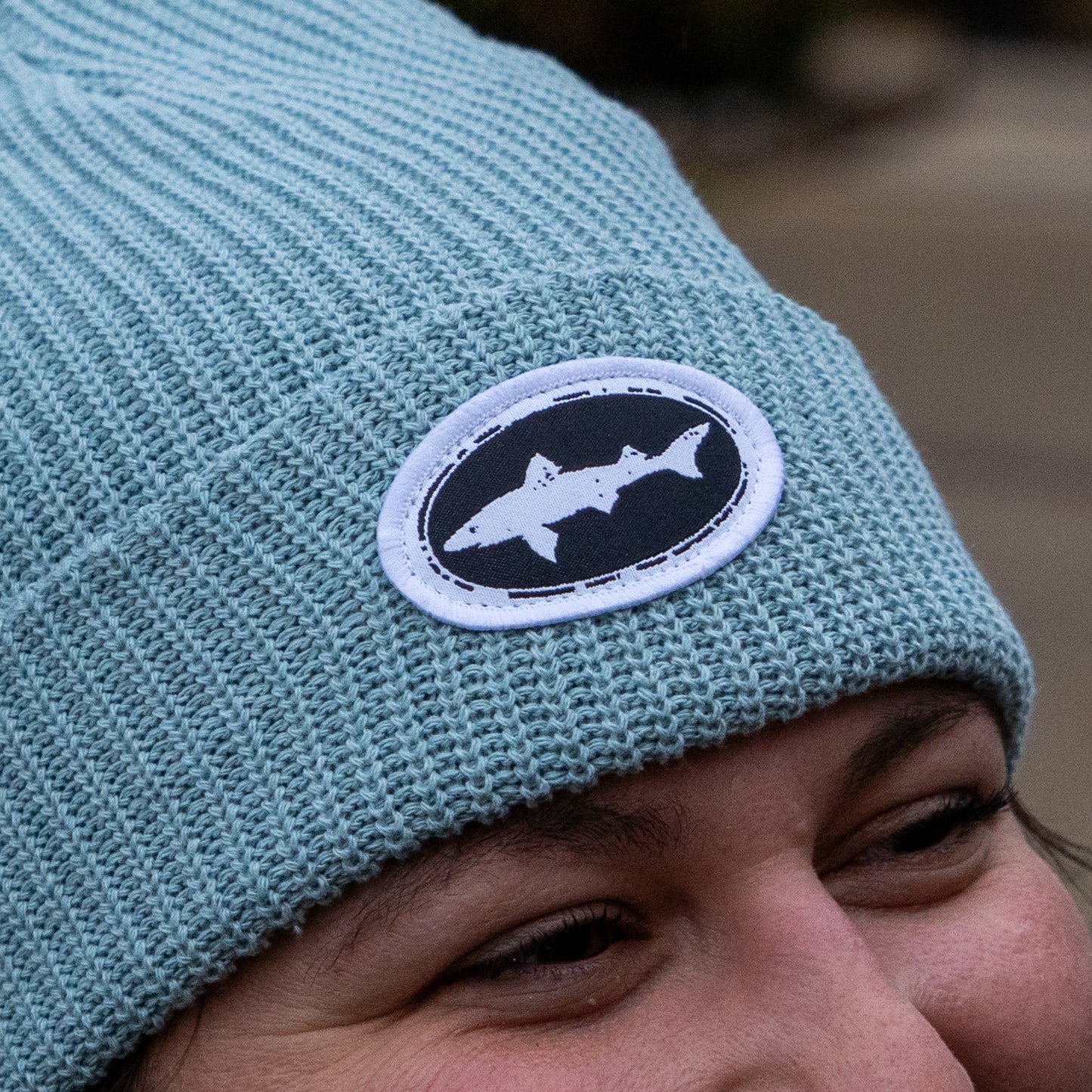 Close-up of a person wearing the Upstate Stock Seafoam Patch Beanie, styled off-center. The beanie is light blue with a white and black woven patch featuring a shark silhouette. Only their eyes and eyebrows are visible.