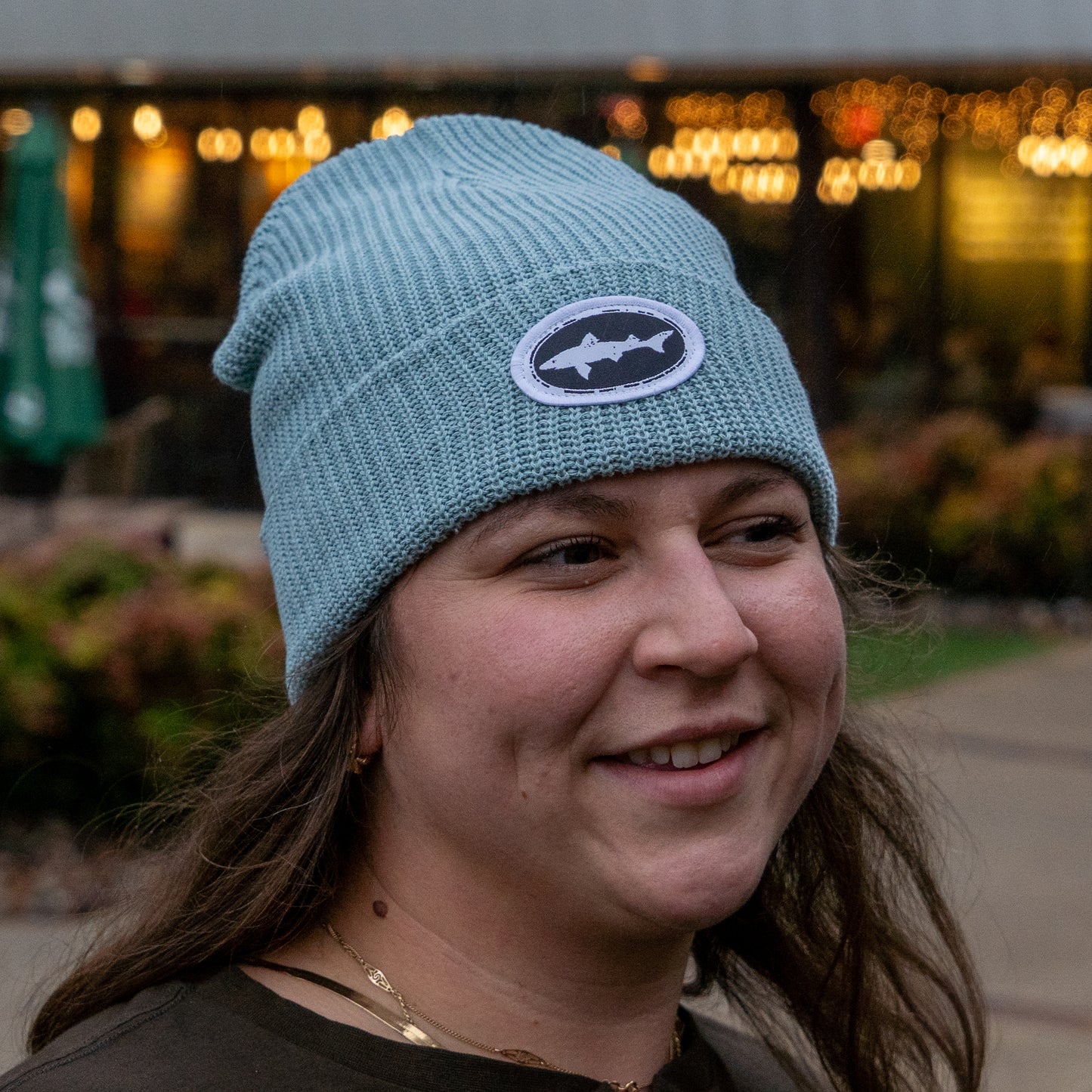 A woman with long brown hair smiles outdoors, wearing the Upstate Stock Seafoam Patch Beanie with an off-center fit and fish emblem. Blurred lights and greenery form the background.