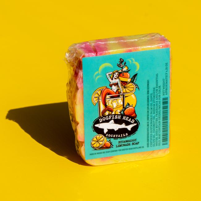 A vibrant Strawberry Lemonade Bar Soap from Indigo Bee rests on a bright yellow surface. The retro label, featuring a woman with fruit and cocktails, reflects the playful spirit of this unique bath product.