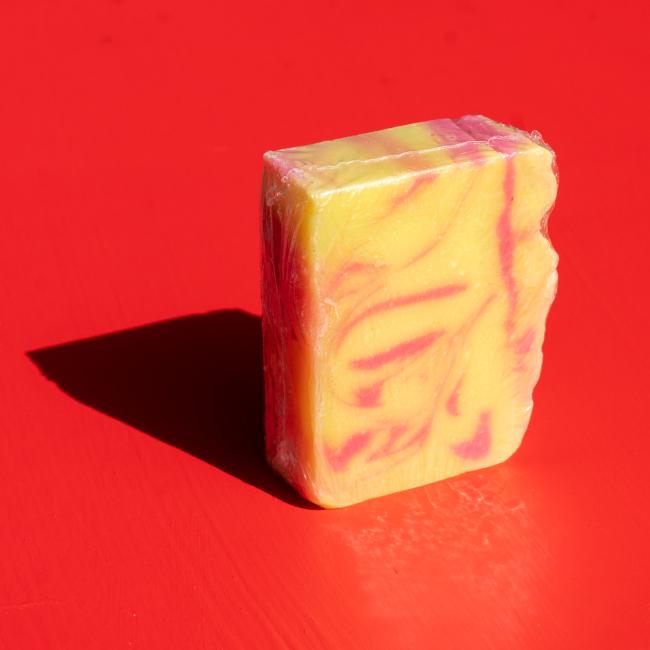 An upright Indigo Bee Strawberry Lemonade Bar Soap, partially wrapped in clear plastic, stands on a bright red surface with its shadow cast to the left.