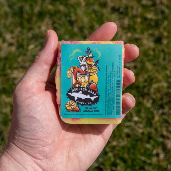 A hand holds an Indigo Bee Strawberry Lemonade Bar Soap with a label showing a dog in a fruit-filled cocktail glass, set against a blurry grass background.