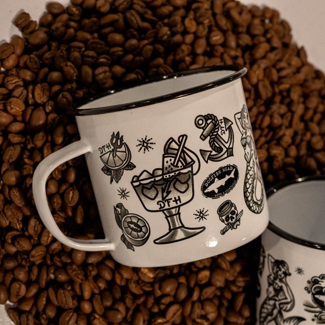 A PROforma Tattoo Enamel Mug, featuring black tattoo-style designs like a cocktail glass, anchor, and skull on white enamel, rests on coffee beans. Another durable Tattoo Enamel Mug with similar art is partially visible nearby.