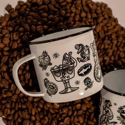 A PROforma Tattoo Enamel Mug, featuring black tattoo-style designs like a cocktail glass, anchor, and skull on white enamel, rests on coffee beans. Another durable Tattoo Enamel Mug with similar art is partially visible nearby.