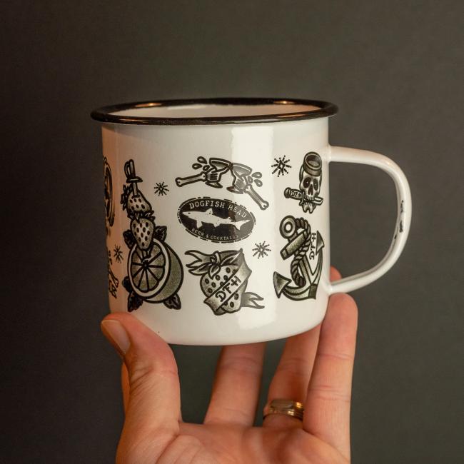 A hand holds the PROforma Tattoo Enamel Mug, its white surface adorned with black tattoo-style art—anchor, lemon, skull, and a Dogfish Head banner. The durable mug stands out against a plain dark background.