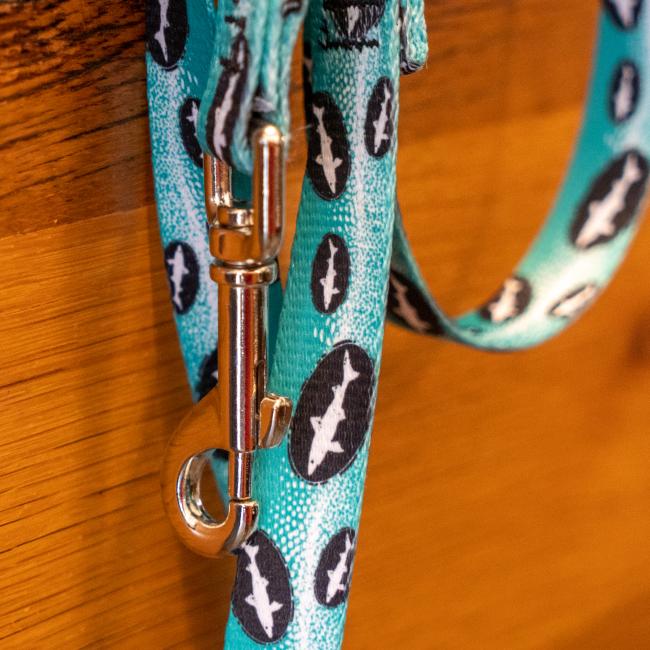 A close-up of the Teal Dog Leash by Lupine, Inc., featuring black and white fish designs, hangs on wood with its metal clasp in view. Pair it with matching collars for a stylish, coordinated look.
