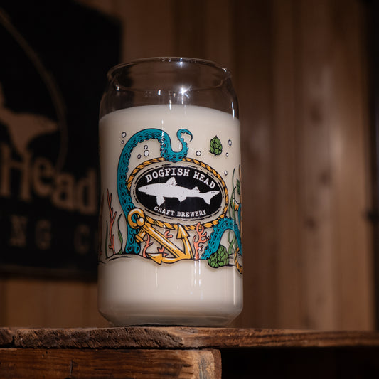 An Under the Sea Can Glass Candle by Unwined Candles, LLC—featuring a Dogfish Head Craft Brewery logo and sea-themed art—sits on a wooden surface, its pale wax evoking the look and scent of the ocean.