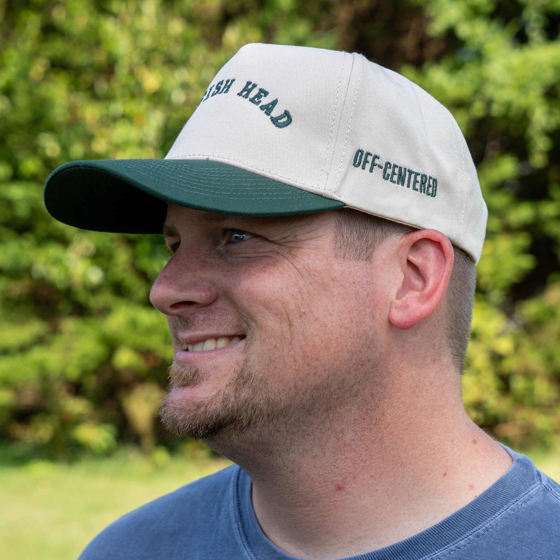 A man with a short beard and mustache smiles outdoors in greenery, wearing a bold Richardson Upright DOGFISH Five Panel Snapback cap in green and white that says FISH HEAD on the front and OFF-CENTERED on the side.