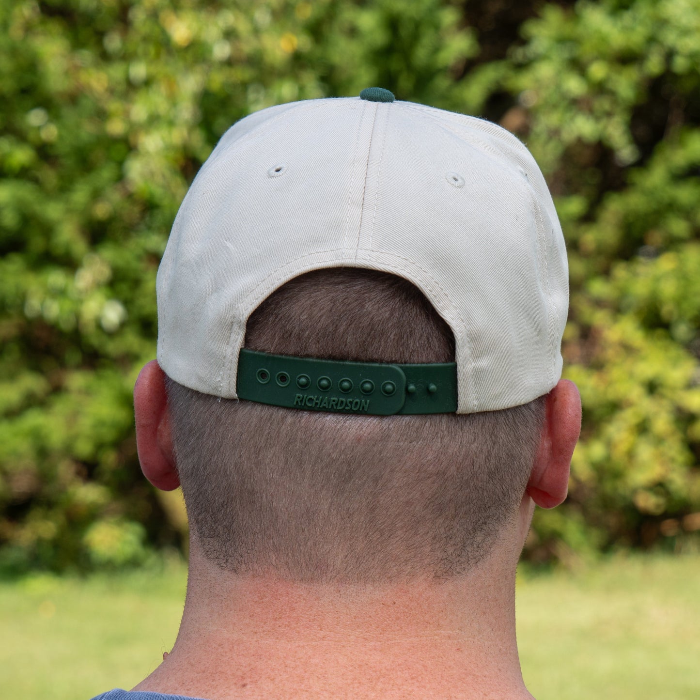Seen from behind amid lush green foliage, a person with short hair sports the Richardson Upright DOGFISH Five Panel Snapback, its adjustable strap visible for bold Dogfish Head-inspired style.