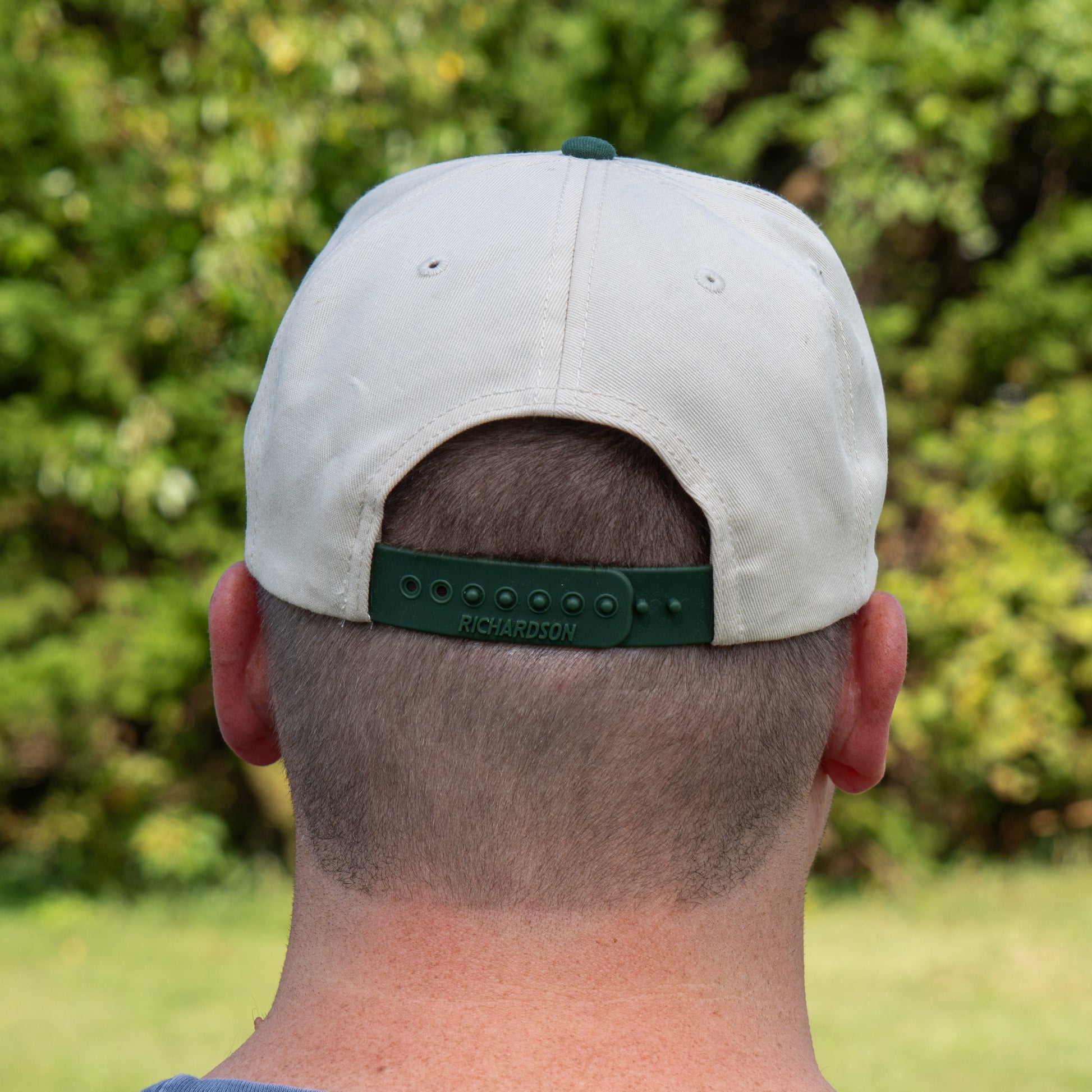 Seen from behind amid lush green foliage, a person with short hair sports the Richardson Upright DOGFISH Five Panel Snapback, its adjustable strap visible for bold Dogfish Head-inspired style.