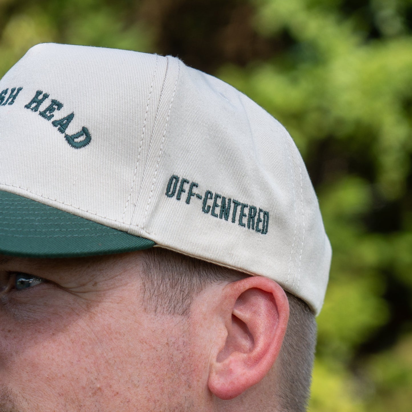 A close-up shows someone in the Upright DOGFISH Five Panel Snapback by Richardson, featuring bold beige and green colors with OFF-CENTERED embroidered on the side. The background is blurred greenery; only part of their face is visible. Inspired by Dogfish Head.