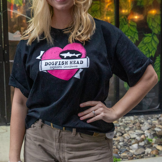 A person stands with one hand on their hip, wearing a GO WEST T-SHIRT COMPANY W Black Tattoo Heart Short Sleeve Tee in soft cotton with a pink heart and white fish logo, paired with tan pants. Rocks and green leaves are in the background.