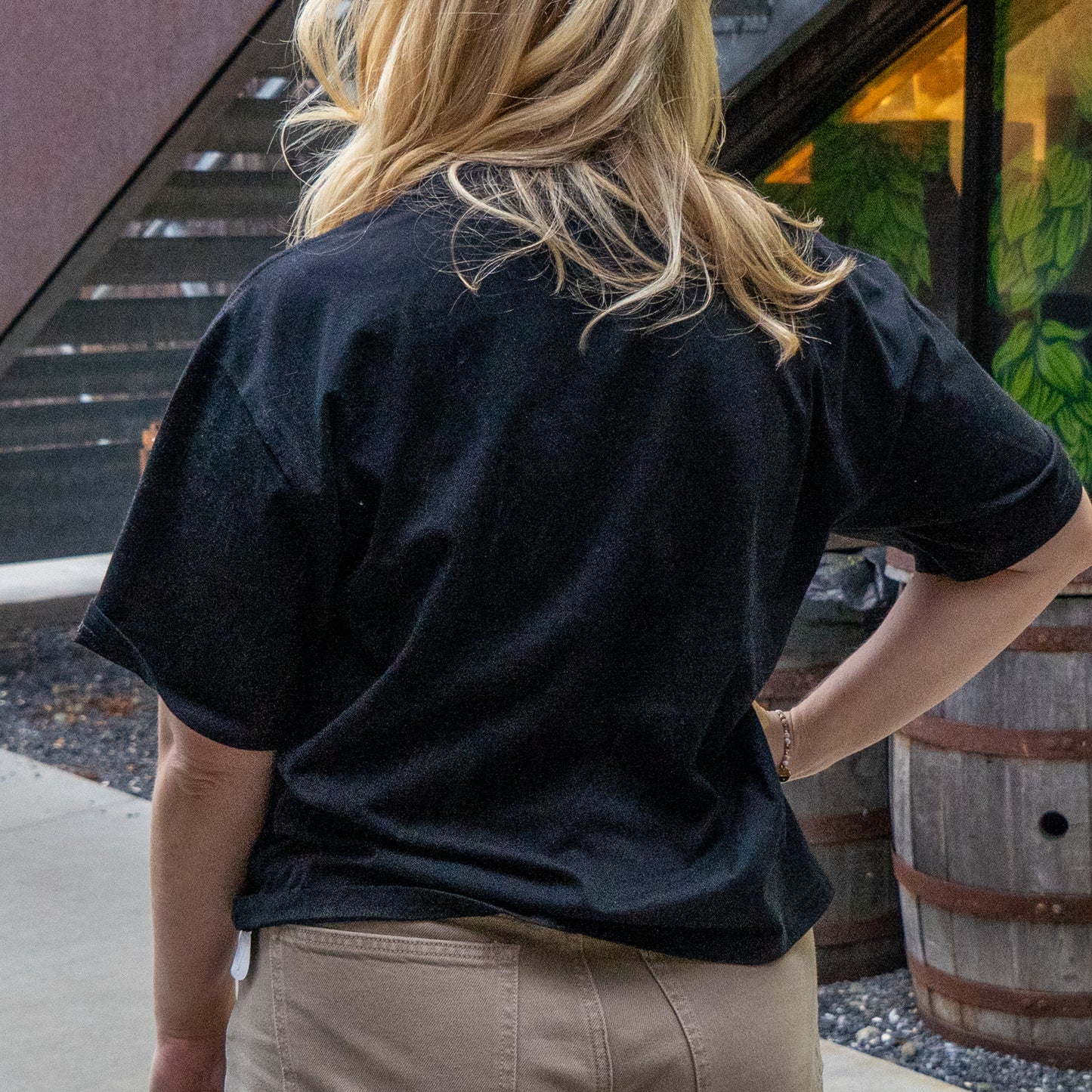 A blonde-haired person stands outdoors with their back to the camera, wearing a soft combed cotton W Black Tattoo Heart Short Sleeve Tee by GO WEST T-SHIRT COMPANY and beige pants near a building and plants.
