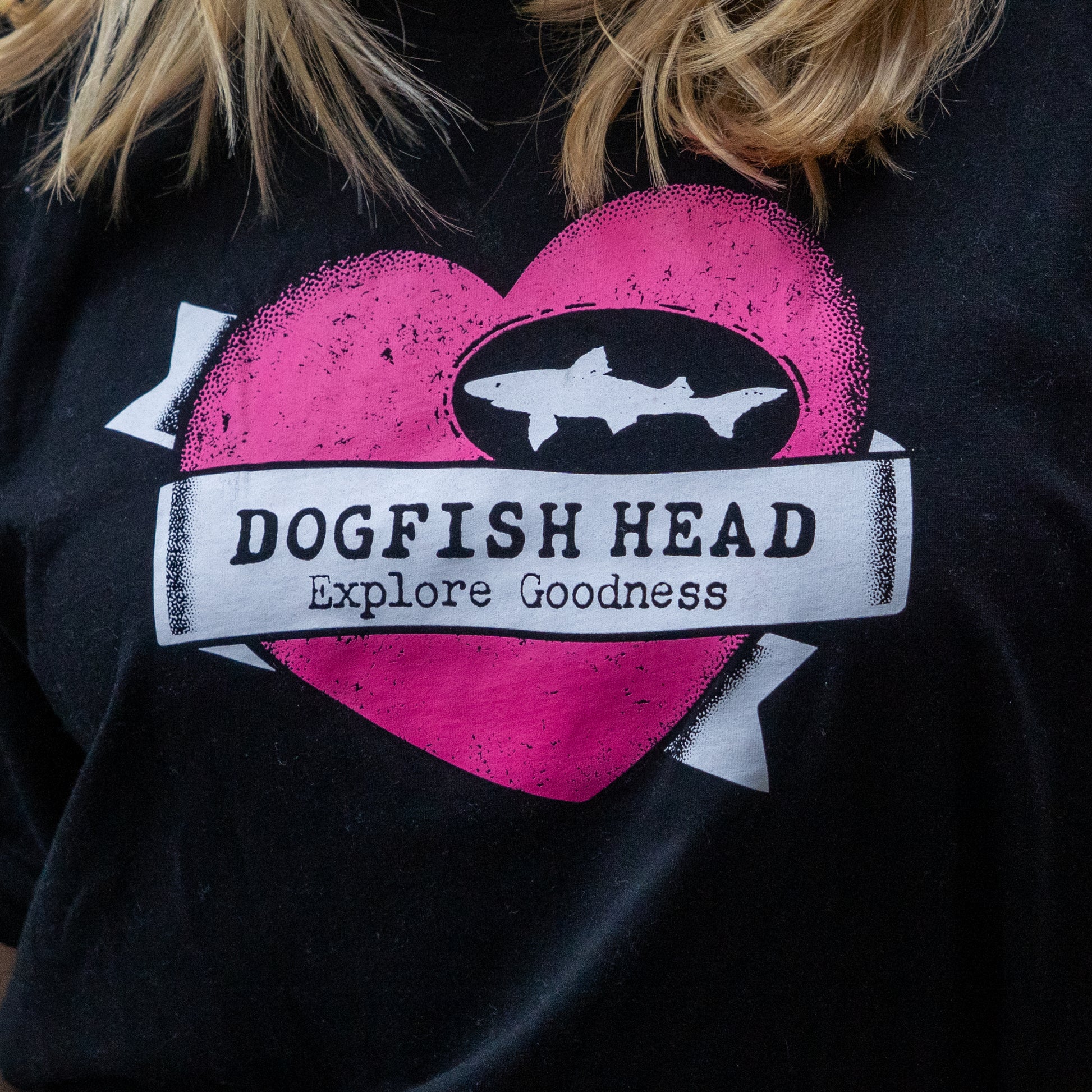 A person wears the GO WEST T-SHIRT COMPANY W Black Tattoo Heart Short Sleeve Tee—crafted from soft combed cotton, with a large pink heart, white dogfish graphic, and "Dogfish Head Explore Goodness" text radiating bold style.