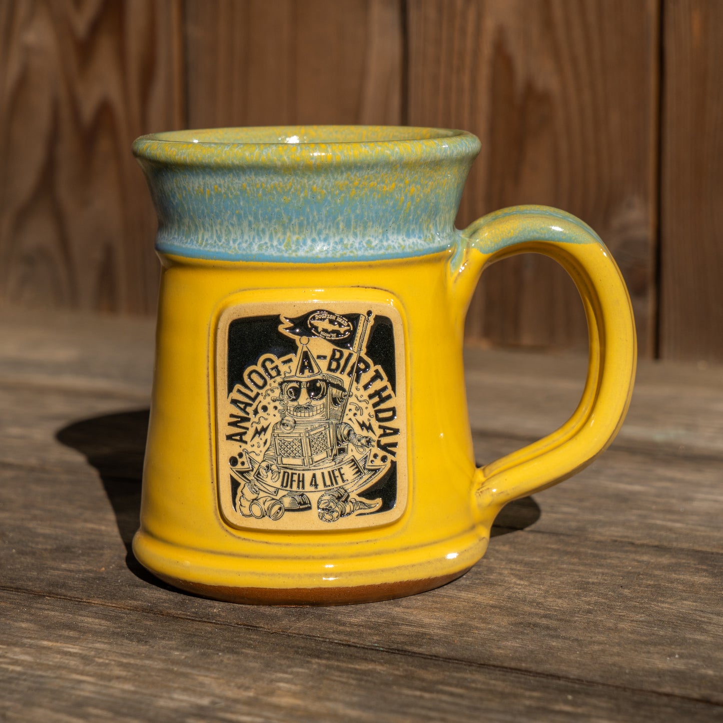 The Deneen Pottery Yellow Analog Splitrock Ceramic Mug, with a blue rim, sits on wood. It displays a robot sticker, the phrases "ANALOG BIRTHDAY" and "FOR A LIFE," plus stars and a cake.
