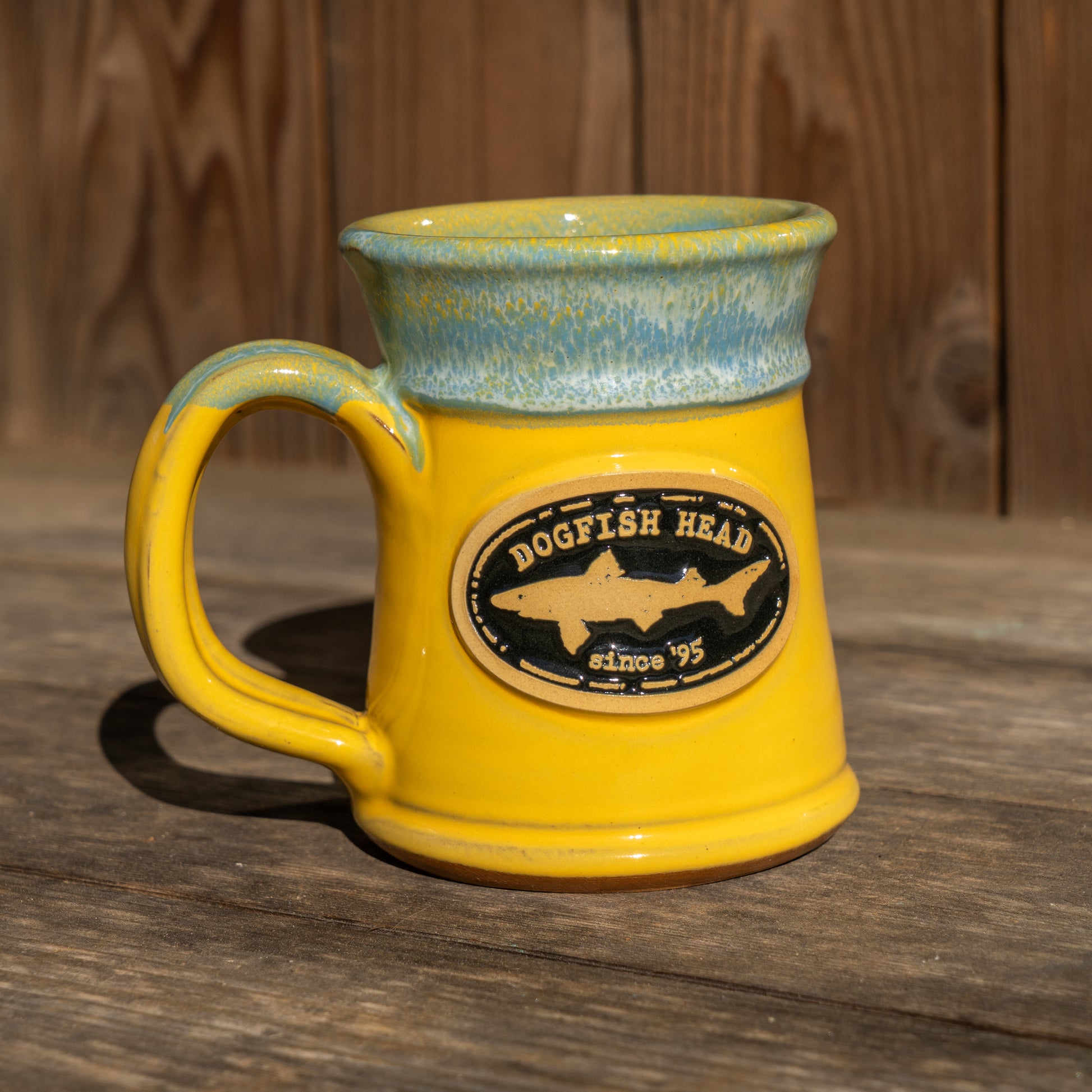 The Yellow Analog Splitrock Ceramic Mug by Deneen Pottery, featuring an oval Dogfish Head since 95 logo and a blue-green rim, sits on a wooden surface with a wooden wall in the background.