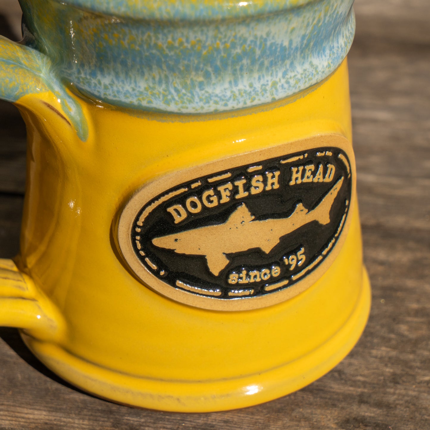 A close-up of the Yellow Analog Splitrock Ceramic Mug by Deneen Pottery, featuring a blue-green rim and an oval logo with "Dogfish Head since 95" and a shark silhouette, resting on a wooden surface.