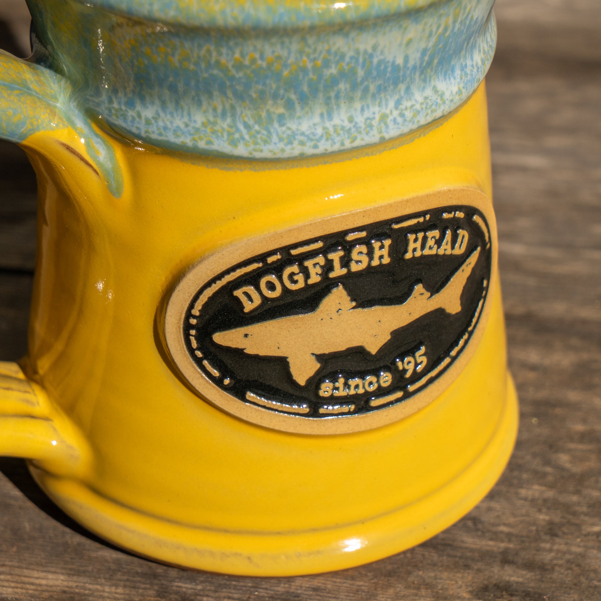 A close-up of the Yellow Analog Splitrock Ceramic Mug by Deneen Pottery, featuring a blue-green rim and an oval logo with "Dogfish Head since 95" and a shark silhouette, resting on a wooden surface.