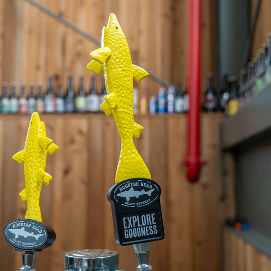 Two neon yellow shark-shaped Merchtank Yellow Shark Tap Handles stand before a wooden wall lined with bottles. One tap displays EXPLORE GOODNESS in bold white text.