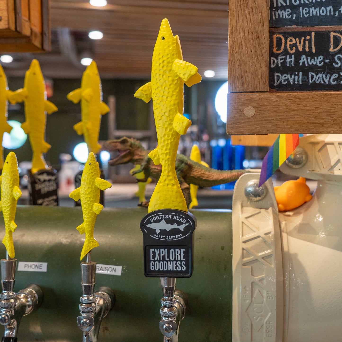 A neon yellow Yellow Shark Tap Handle by Merchtank, shaped like a fish, is pictured. Other tap handles, a toy dinosaur, and part of a rainbow flag are visible in the background.